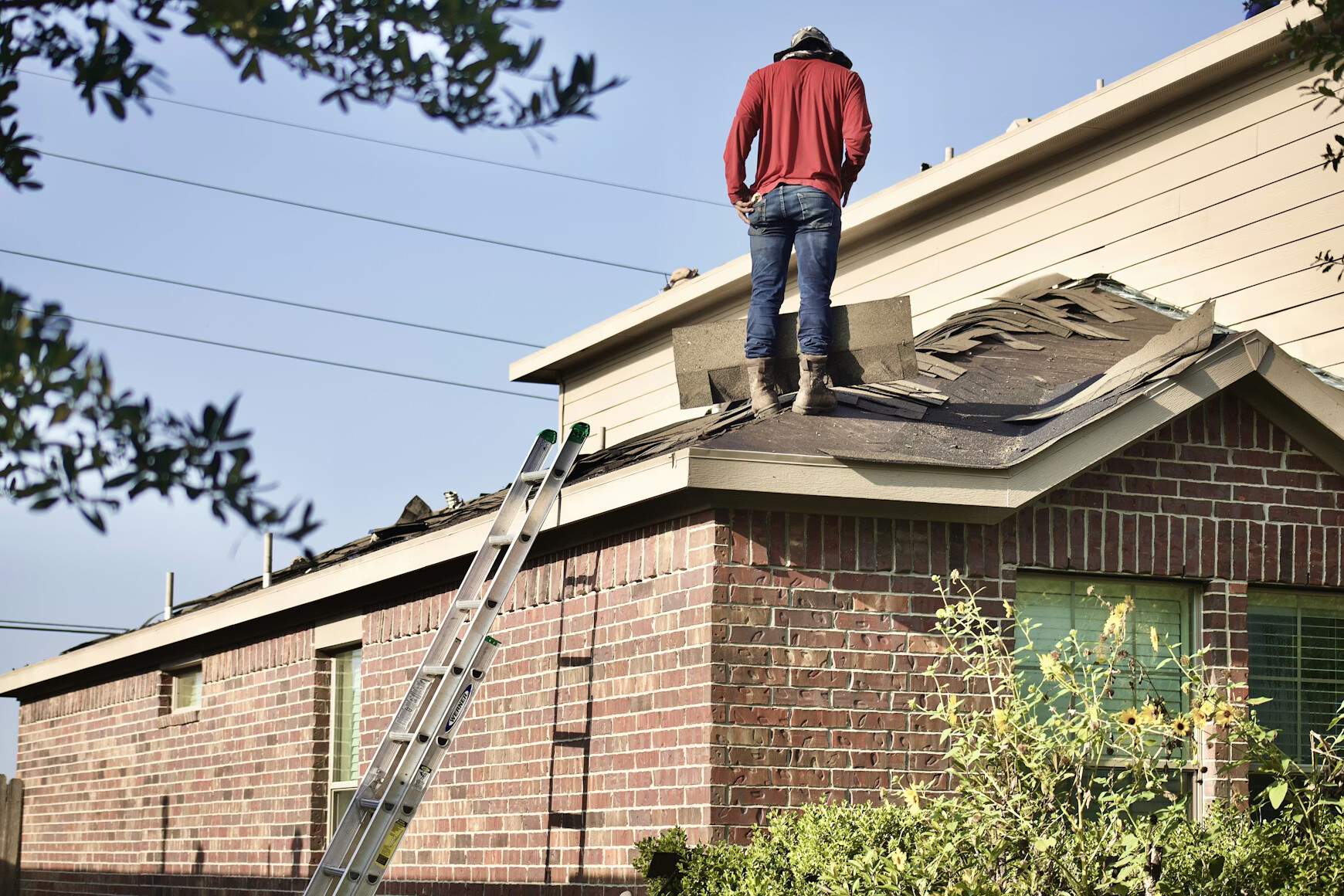 4c9d2398-b4cd-4678-ba57-a1dc95b66edd A construction worker wearing a yellow hard hat, safety goggles, and an orange high-visibility vest uses a power drill to secure grey roof tiles on a bright, sunny day.